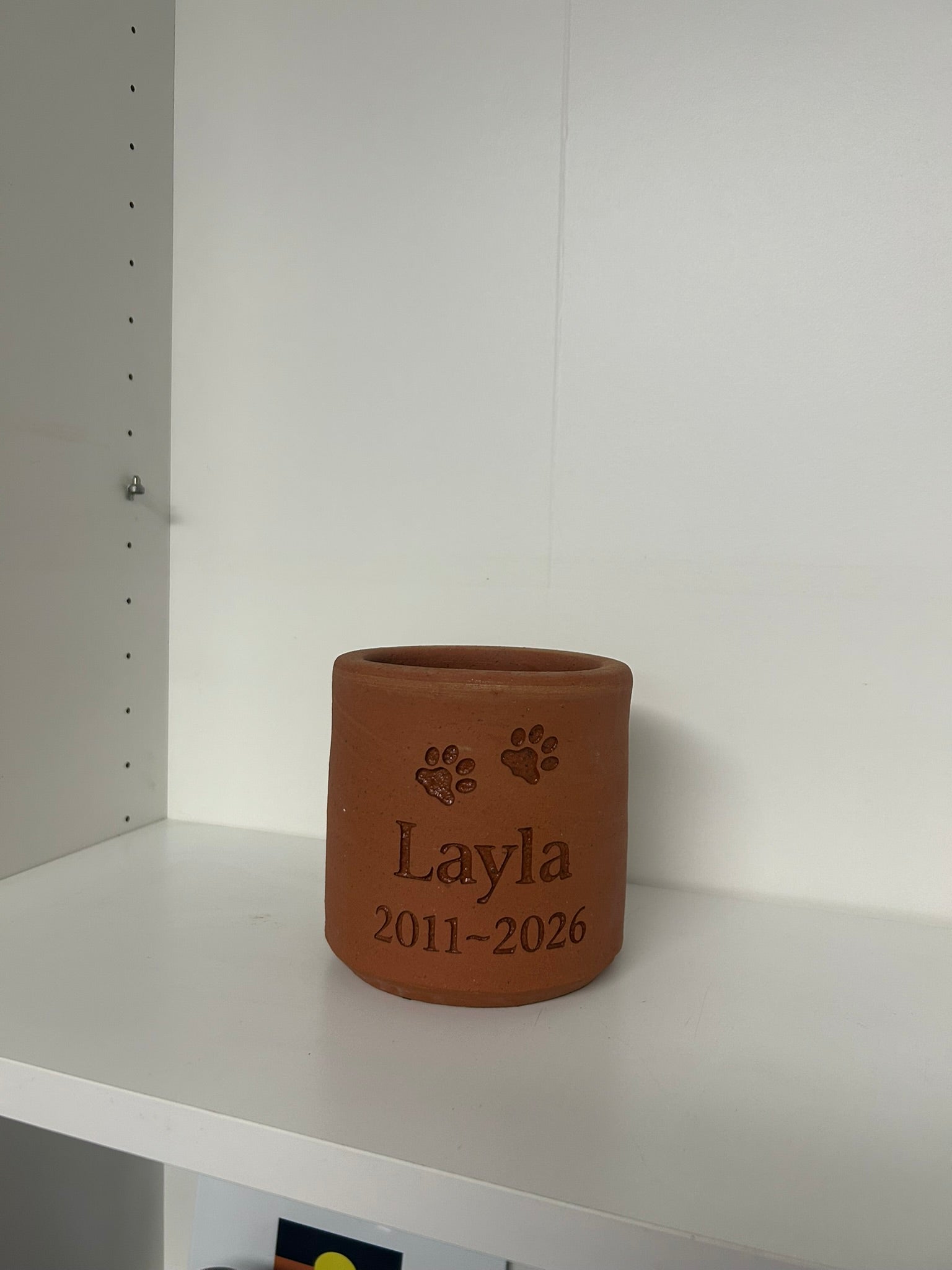 Brown ceramic pot with engraved name 'Layla' and years '2011-2026' on a white shelf.